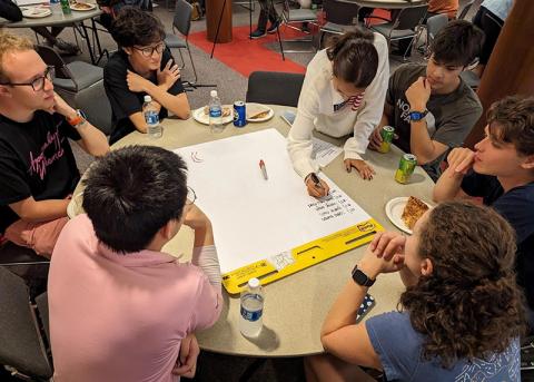 A group of students work at a round table while one student writes on a large pad of paper
