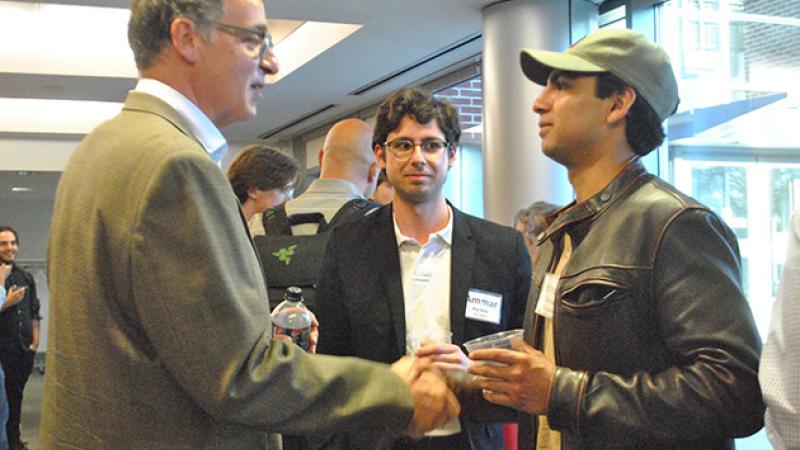 Student in a baseball hat shakes hands with a man in a suit while a third man looks on