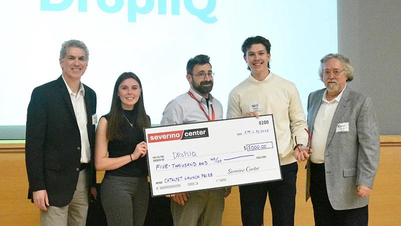 Students and advisors pose in front of logo and hold giant check