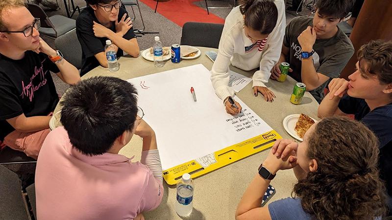 A group of students work at a round table while one student writes on a large pad of paper