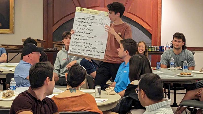 Student stands in front of groups of students presenting content from a large pad of paper